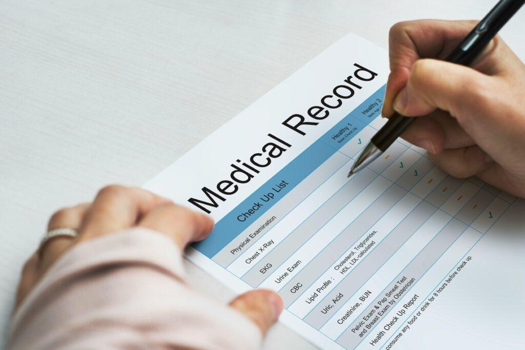 Close-up of hands holding a pen and filling out a printed medical record checklist on a desk, with the heading “Medical Record” visible and rows of health examinations and test results marked on the form.