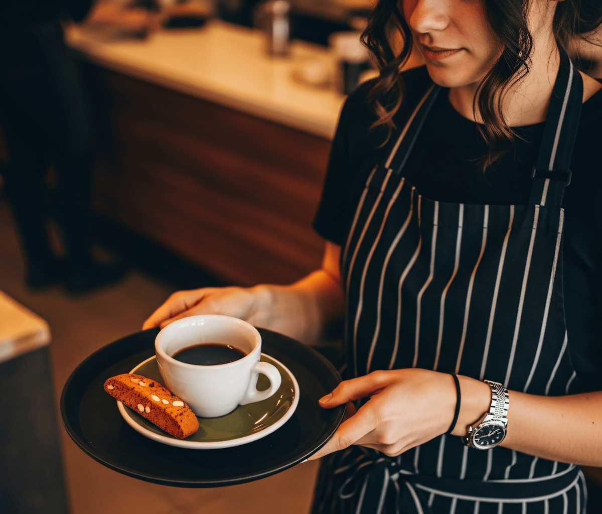 A woman in an apron holds a plate with a cup of coffee, smiling as she prepares to serve.