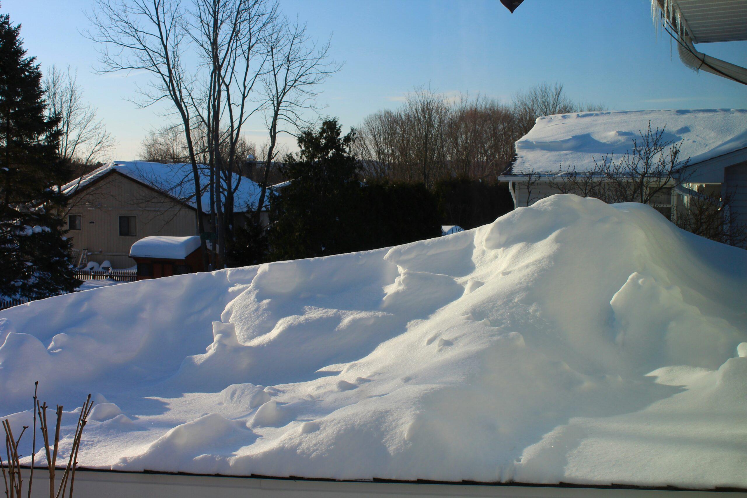 Large accumulation of snow piled high on a rooftop in a residential neighborhood, with nearby houses, leafless trees, and a clear blue winter sky in the background